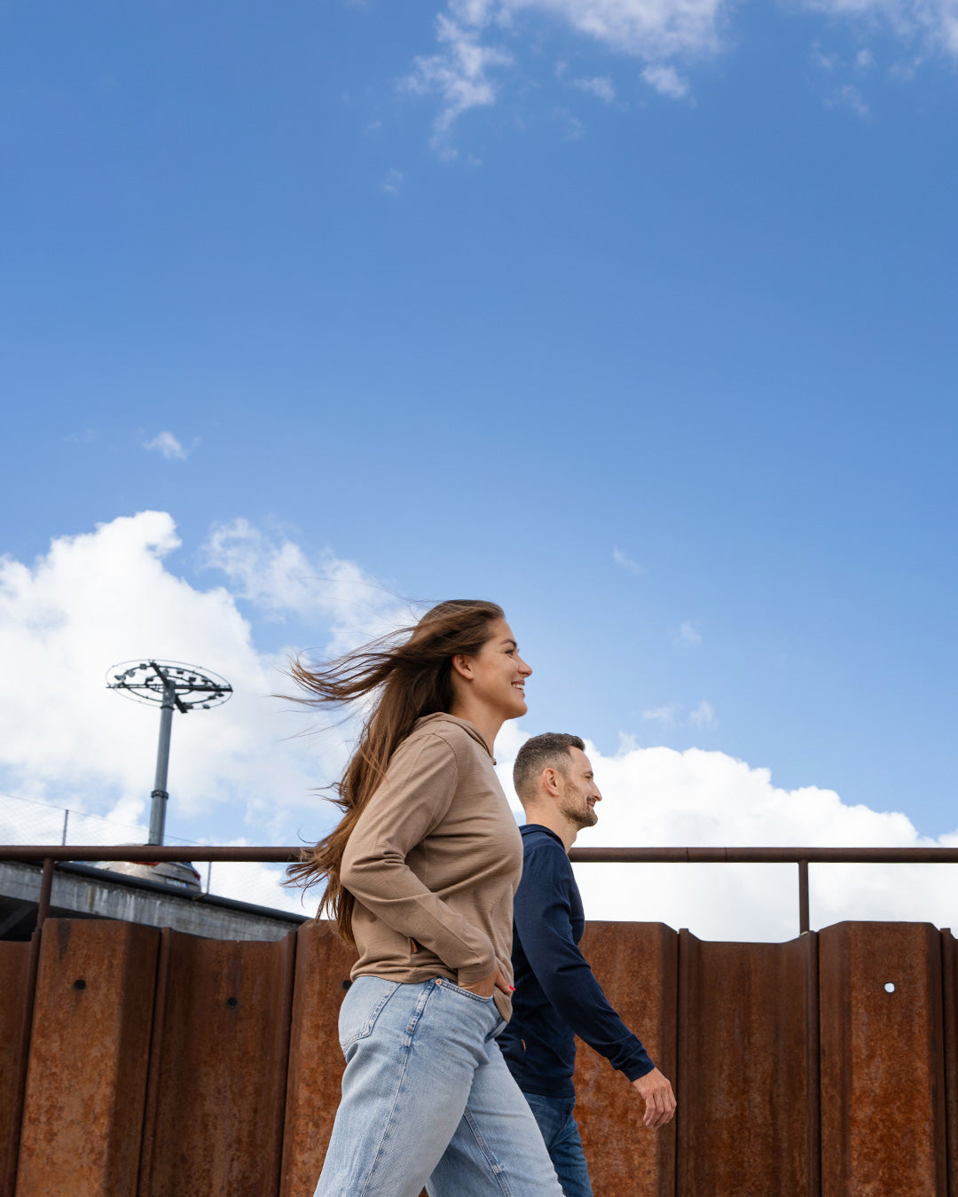 A smiling woman with long brown hair blowing in the wind walks outdoors alongside a man. She is wearing a tan hoodie and light blue jeans, while he is wearing a dark blue long-sleeved shirt. They are walking past a rusted corrugated metal wall under a bright blue sky with scattered white clouds.
