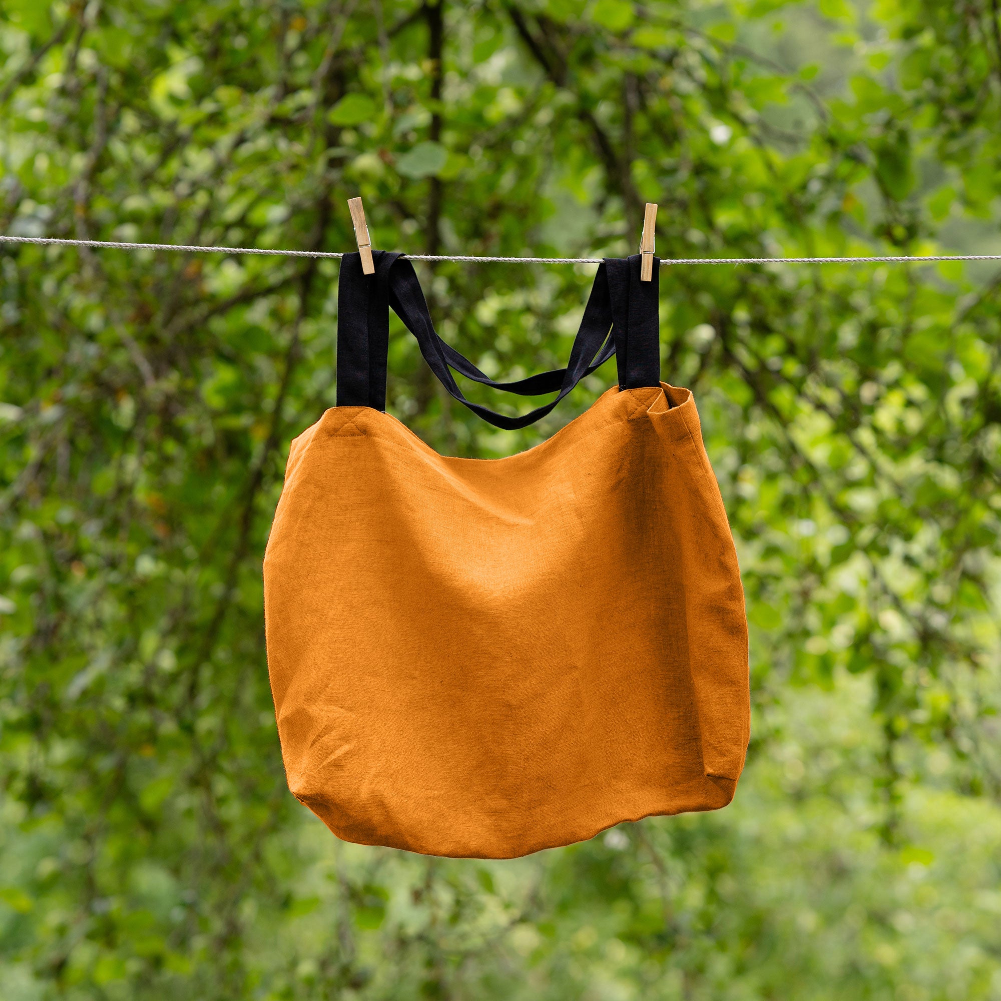 The menique linen tote bag in stone green color with black handles hung on a clothesline outside.