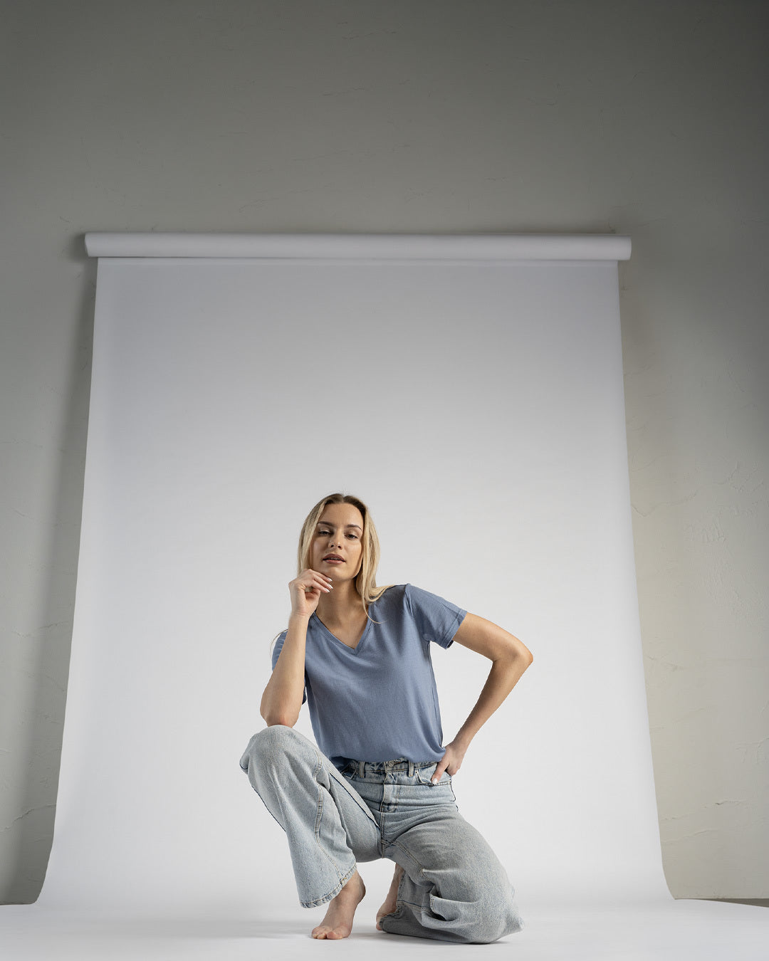 A woman with blonde hair sitting on a stool in a studio against a white seamless backdrop, wearing a blue t-shirt and light blue jeans with one hand resting near her chin.