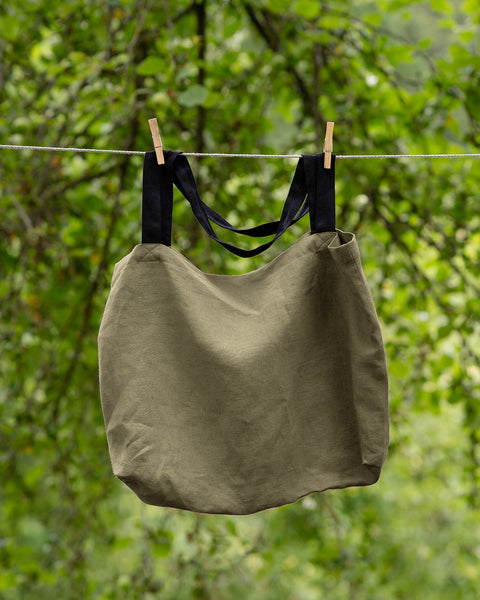 The menique linen tote bag in stone green color with black handles hung on a clothesline outside.