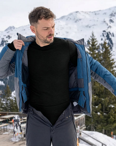 Man wearing a black merino wool thermal base layer while putting on a ski jacket in snowy mountains, demonstrating slim-fit comfort and moisture-regulating winter layering.