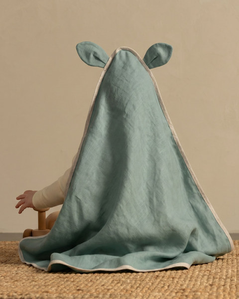 A baby sitting on a woven natural fiber rug, viewed from behind, completely covered by a sage green hooded baby towel. The hood of the towel features two small, cute animal-like ears. The towel has a light beige binding along its edges. One small hand reaches out towards a wooden toy out of focus in the foreground. The background is a solid, warm taupe wall.