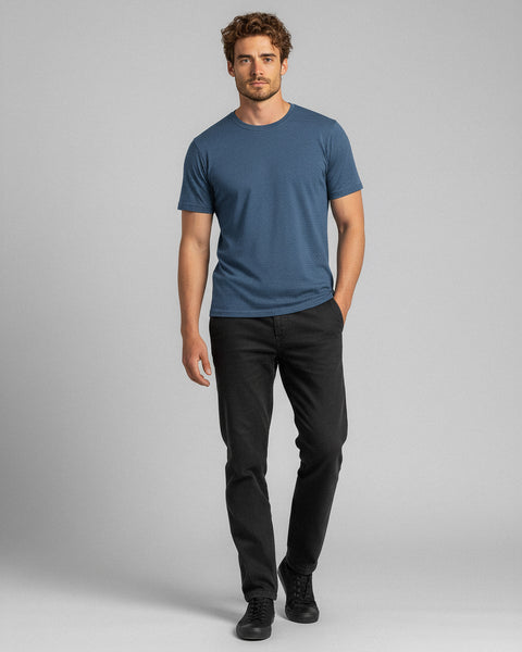 A man walks forward wearing a blue T-shirt, black jeans, and black sneakers, standing against a plain gray background.