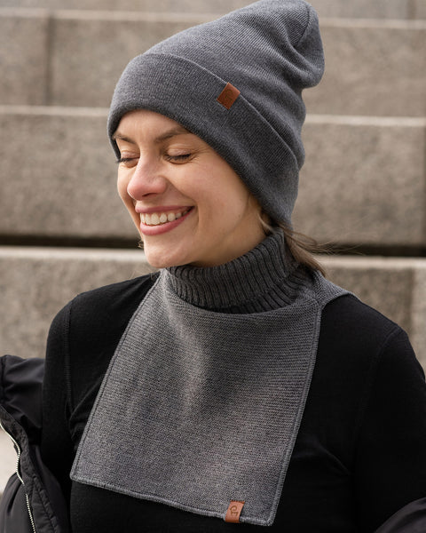 A woman is shown smiling brightly with her eyes closed, facing slightly to her left. She is wearing a dark grey knit beanie with a small brown logo patch on the cuff. She's also wearing a matching dark grey knit neck warmer, featuring a ribbed turtleneck and a flat-knit bib extending down her chest, also with a small brown logo patch at the bottom corner. Similar to the first image, she has a black long-sleeved shirt on and a dark jacket slipping off her shoulders.