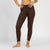 A person from the waist down stands in profile, facing left, against a plain white background. They are barefoot and wearing fitted, brown leggings, with their hands resting on their hips.