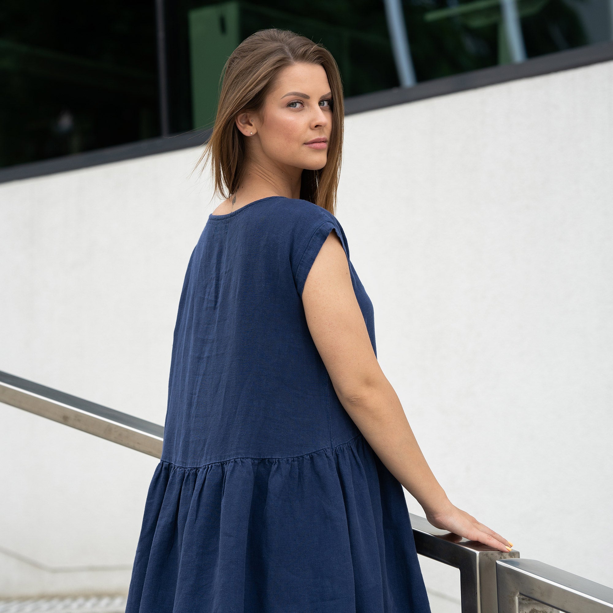 Young woman posing in a city street on stears wearing linen smock dress cecilia in a storm blue color. Girl with a cat tattoo on her left arm.