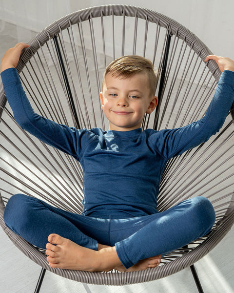 A boy sitting in a lounge chair, wearing the menique Kids&#39; Merino 160 Long Sleeve Set in Denim color. 